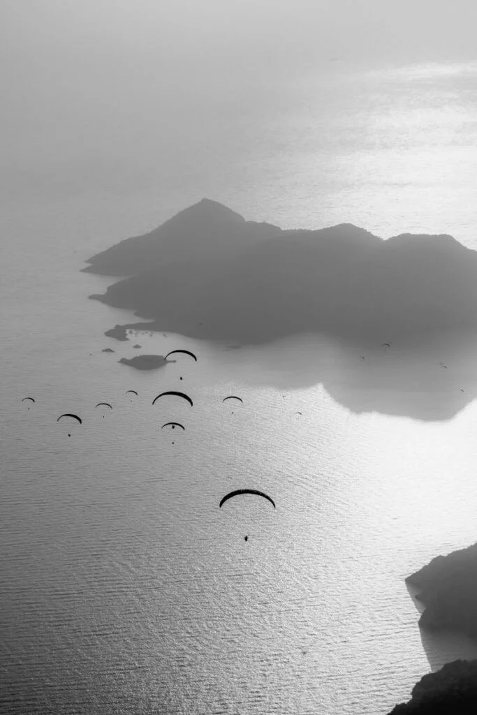 A dramatic black and white aerial photo of paragliders soaring above misty islands and the ocean.